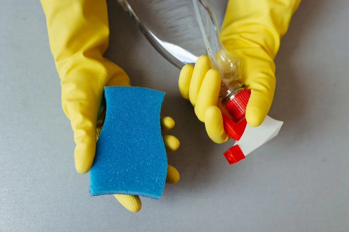 Mãos com luvas de proteção a segurar uma esponja e um frasco de produto de limpeza.